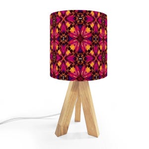 May include: A small table lamp with a wooden tripod base and a cylindrical lampshade. The lampshade features a vibrant, colorful pattern in shades of pink, orange, and purple. The lamp is designed for indoor use.