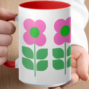 A person's hand holding a white mug with a red interior and a design of two pink flowers with green leaves.