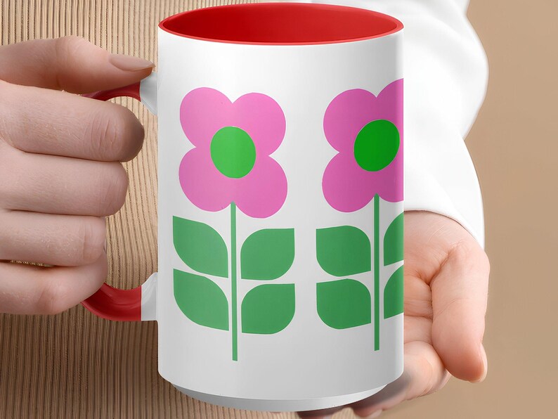 A person's hand holding a white mug with a red interior and a design of two pink flowers with green leaves.