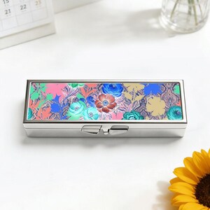 May include: A rectangular, silver pill box with a floral design on the lid. The lid features a colourful pattern of flowers in shades of pink, blue, and green. The box has a hinged closure.
