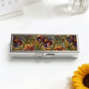 May include: A rectangular, silver-colored pill box with a hinged lid. The top of the box features a colorful floral design in shades of purple, orange, yellow, and green, resembling stained glass. The box is closed and sits on a white surface.