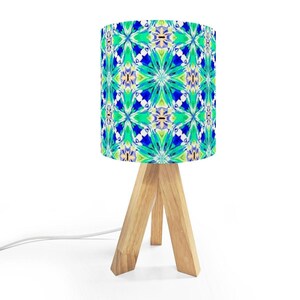 May include: A small table lamp with a cylindrical lampshade featuring a vibrant, abstract pattern in shades of blue, green, and yellow. The lampshade sits atop a natural wood tripod base, with a white electrical cord extending from the base.