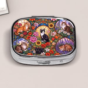 May include: An oval metal pill box featuring a colorful stained glass style illustration of five cats surrounded by various flowers including sunflowers, poppies, and lilies. The cats are depicted in different poses and colors, including orange tabby, black and white, and brown tabby, set against a vibrant, intricate floral background.