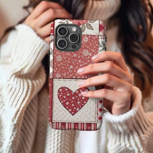 Può includere: Una custodia per telefono con un motivo floreale rosso, bianco e floreale, con un design a forma di cuore. La custodia è progettata per un iPhone e ha un ritaglio per l'obiettivo della fotocamera.
