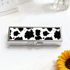 May include: A rectangular, silver pill box with a hinged lid. The top of the box has a black and white cow print design. The box is on a white surface, with a sunflower in the lower right corner.