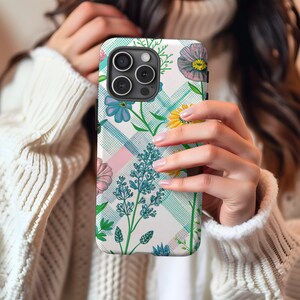 Può includere: Una custodia per telefono bianca con un motivo floreale. La custodia ha un design floreale blu, rosa e giallo su sfondo bianco. La custodia ha un motivo a scacchi sullo sfondo.