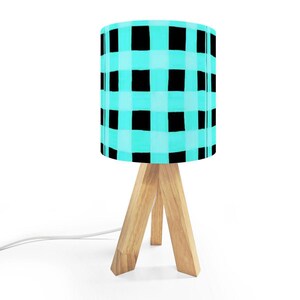May include: A small table lamp with a light wood tripod base and a cylindrical lampshade. The lampshade features a turquoise and black checkered pattern. The lamp is plugged into a white electrical cord.