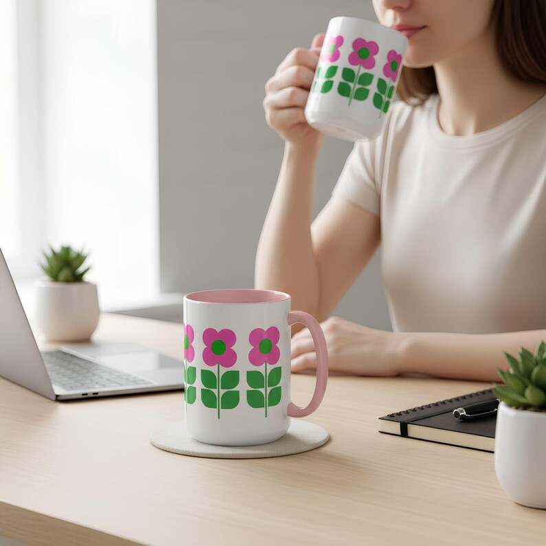 A woman is sitting at a desk, drinking from a mug with a floral design, while a laptop and a notebook are visible on the desk.