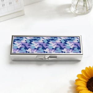 May include: A rectangular, silver pill box featuring a blue and purple camouflage pattern. The box has a hinged lid and a clasp. The background is white. The box is approximately 10 cm long.
