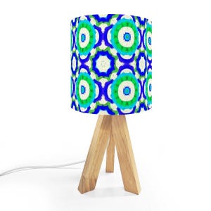May include: A small table lamp with a cylindrical lampshade featuring a repeating pattern of blue, green, and white circular designs. The lamp has a natural wood tripod base and a white power cord.