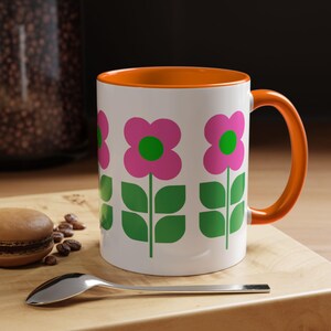 A white mug with a floral design and an orange handle, placed on a wooden surface next to a spoon and some coffee beans.