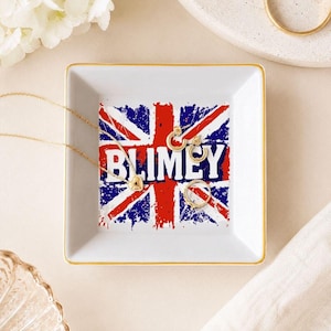 May include: A square white ceramic trinket dish with a gold-toned rim. The center features a distressed Union Jack flag graphic in red, white, and blue with the word BLIMEY printed in bold white capital letters across the middle of the design.