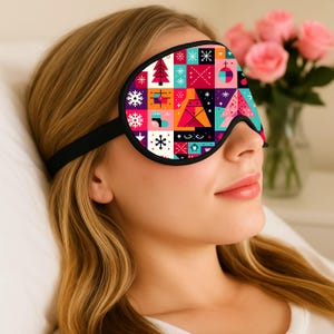 May include: A black sleep mask with a colorful geometric pattern featuring Christmas trees, snowflakes, and other festive shapes. The mask has a black strap and is worn over the eyes. The background includes a bouquet of pink roses.