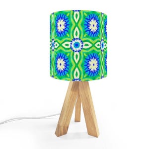 May include: A small table lamp with a cylindrical shade featuring a vibrant green, blue, and white floral pattern. The lamp has a natural wood tripod base and a white power cord.