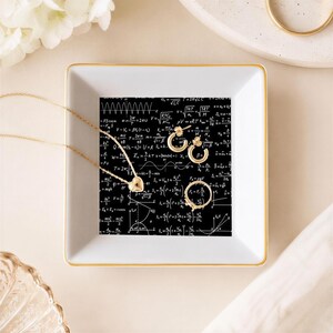 May include: A square white ceramic trinket dish with a gold-toned rim, featuring a black interior base printed with white mathematical formulas and physics equations. A gold heart-shaped pendant necklace and two small gold hoop earrings rest inside.