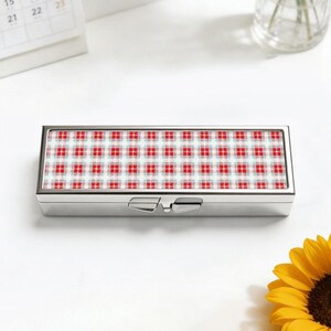 May include: A rectangular, silver pill box featuring a red, white, and grey plaid design on the lid. The box is closed and has a hinged clasp. The background is white, with a calendar and a sunflower visible.