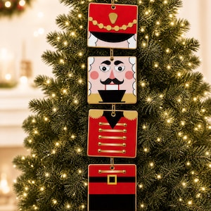 May include: A vertical hanging Christmas ornament consisting of four square panels connected by gold chains. The panels feature a red nutcracker design with a hat, a face with a mustache, a red jacket with gold buttons, and a black belt with a gold buckle.
