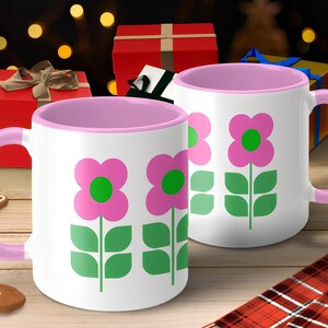 A pair of pink and white mugs with green flower designs, placed on a wooden surface with a gingerbread cookie and wrapped gifts in the background.