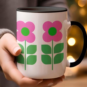 A person's hand holding a white mug with a floral design in pink and green colors.