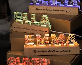May include: Four custom name signs made of clear resin embedded with dried flowers, mounted on rectangular wooden bases. Each sign features LED lighting and engraved text, including phrases like May love and happiness follow you wherever you go.
