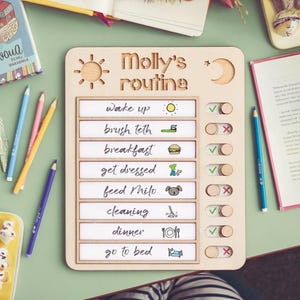 Op de afbeelding: Houten dagelijkse routinekaart met de tekst "Molly's routine". De kaart bevat secties voor wakker worden, tanden poetsen, ontbijt, aankleden, Milo voeren, schoonmaken, dineren en naar bed gaan. Een zon en maan zijn uitgesneden.