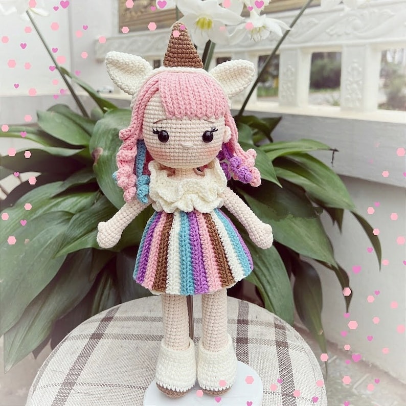Puede incluir: Una mu&ntilde;eca de crochet con un vestido rosa y morado, un cuerno de unicornio y un sombrero de cono de helado blanco.