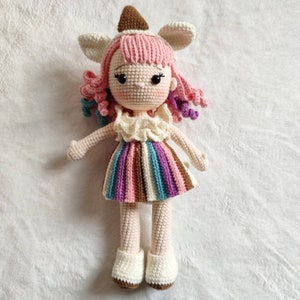 Puede incluir: Una mu&ntilde;eca de crochet con pelo rosa, un vestido a rayas y zapatos blancos. La mu&ntilde;eca lleva un gorro de crochet marr&oacute;n y blanco y un cuello de crochet blanco.