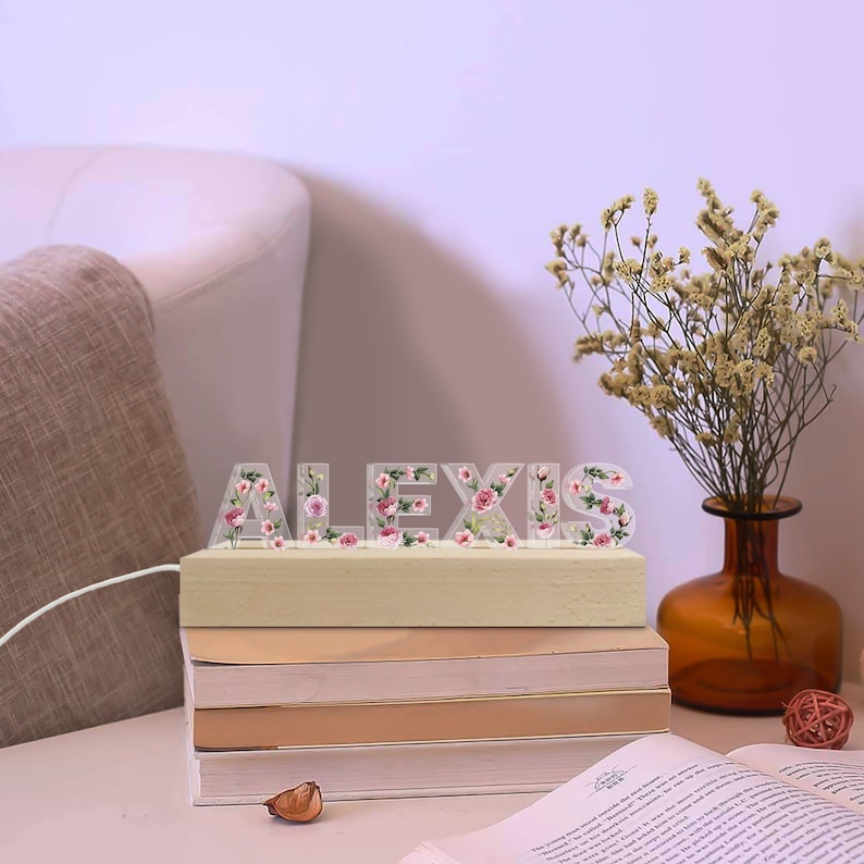 May include: A decorative light spelling "ALEXIS" with clear letters filled with pink and red flowers. The letters sit on a wooden base, illuminated by a soft glow. The lamp is placed on a stack of books, with a vase of dried flowers in the background.