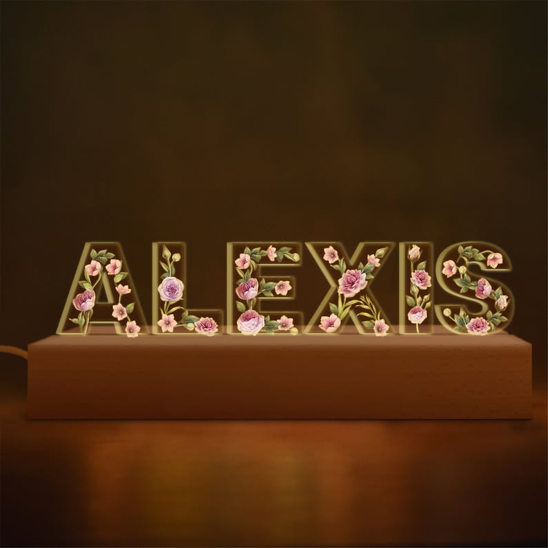 May include: A decorative light spelling "ALEXIS" with floral accents. The letters are illuminated and sit on a wooden base. The flowers are pink and purple, adding a touch of elegance to the design. The background is a warm, dark brown.