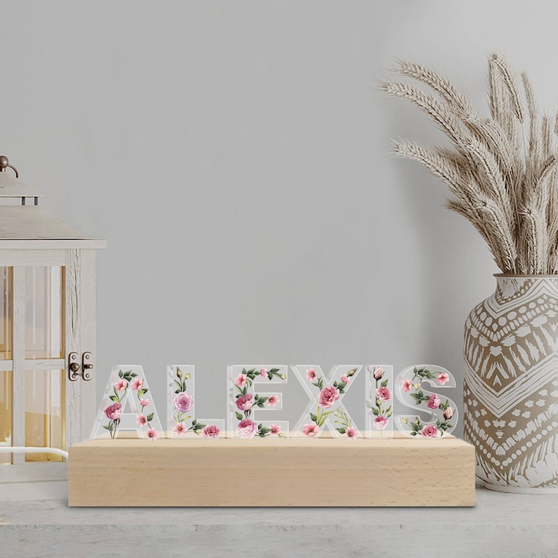 May include: A wooden base supports the name "ALEXIS" spelled out in clear acrylic letters. Each letter is adorned with a floral design featuring pink and red roses and green leaves. The letters are illuminated, creating a decorative display.