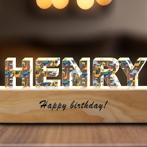 Może przedstawiać: Drewniana podstawa z wygrawerowanym napisem "Happy birthday!" Na podstawie znajdują się pięć akrylowych liter tworzących imię "HENRY". Litery są ozdobione kolorowym wzorem z dinozaurami.