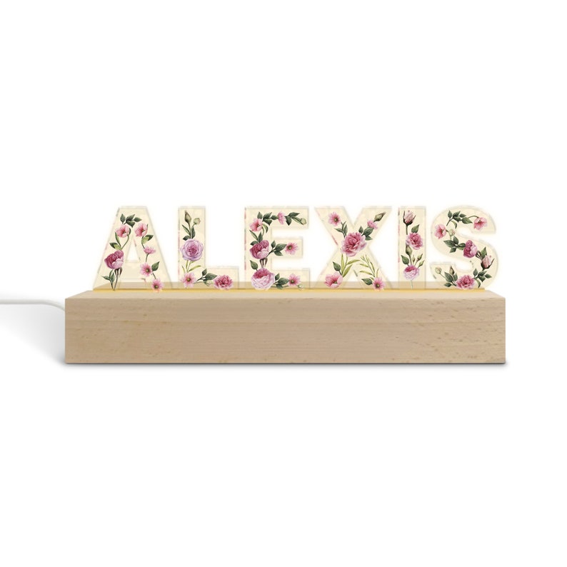 May include: A wooden light-up sign spelling "ALEXIS" with floral designs. The letters are cream-colored and adorned with pink and purple flowers and green leaves. The sign sits on a light-colored wooden base and has a white power cord.