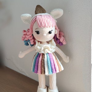 Puede incluir: Una mu&ntilde;eca de crochet con pelo rosa, un vestido blanco con rayas de colores y zapatos blancos. La mu&ntilde;eca lleva un gorro de crochet marr&oacute;n y blanco.
