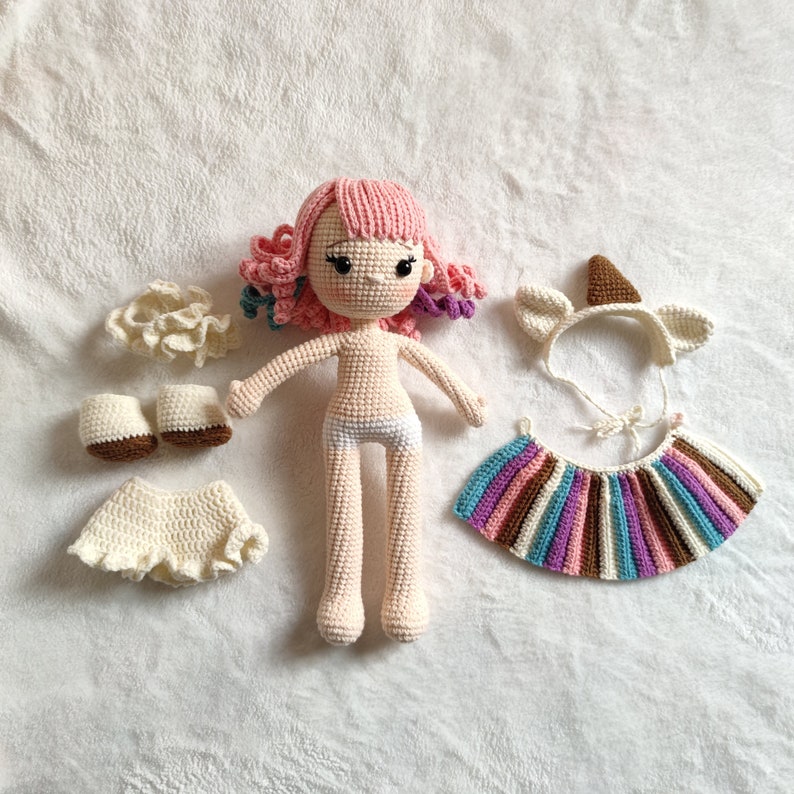 Puede incluir: Una mu&ntilde;eca de crochet con pelo rosa, que lleva un pa&ntilde;al blanco y una variedad de accesorios de ropa de crochet, incluyendo una falda, zapatos, una diadema y una falda de rayas de colores.