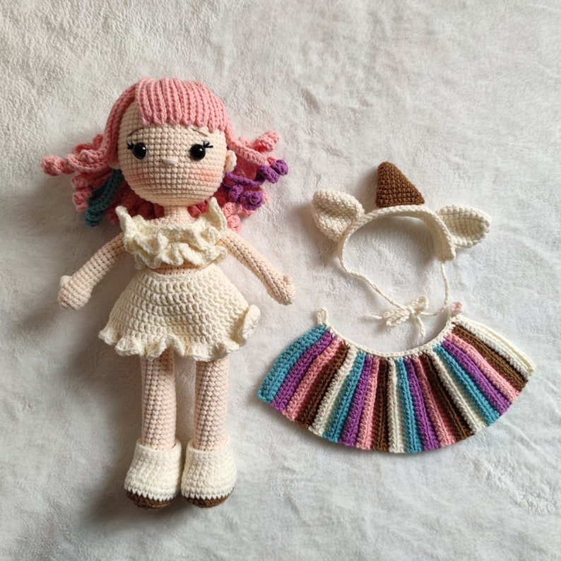 Puede incluir: Una mu&ntilde;eca de crochet con pelo rosa, un vestido blanco y una falda blanca de crochet. La mu&ntilde;eca lleva una diadema blanca de crochet con un cuerno marr&oacute;n de crochet. La mu&ntilde;eca est&aacute; sentada sobre una superficie blanca.