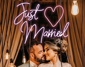 May include: A newly married couple poses for a photo with a pink neon sign that reads "Just Married" with a heart in the middle. The couple is standing in front of a backdrop of brown curtains and string lights.