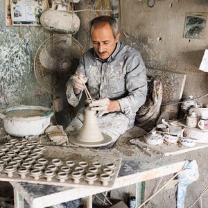 Può includere: Un uomo in un laboratorio polveroso sta modellando un vaso di terracotta su un tornio da vasaio. Sta usando uno strumento per lisciare l'argilla. Ci sono molti altri vasi di terracotta su un tavolo sullo sfondo.