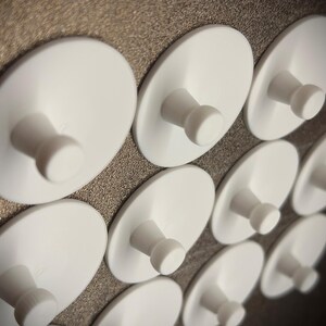 Könnte beinhalten: Nahaufnahme einer Wand, die mit weißen, runden, selbstklebenden Haken bedeckt ist. Die Haken haben einen kleinen, runden Knopf in der Mitte.