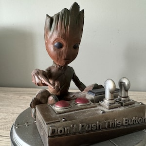 May include: A brown and green figurine of a character from the Guardians of the Galaxy movie series, Groot, sitting on a silver base with a control panel. The control panel has two red buttons and a sign that reads "Don't Push This Button".