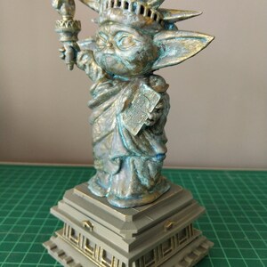 May include: A green and gold statue of the Statue of Liberty, but with the head and body of a Baby Yoda character. The statue is standing on a square base with decorative details.