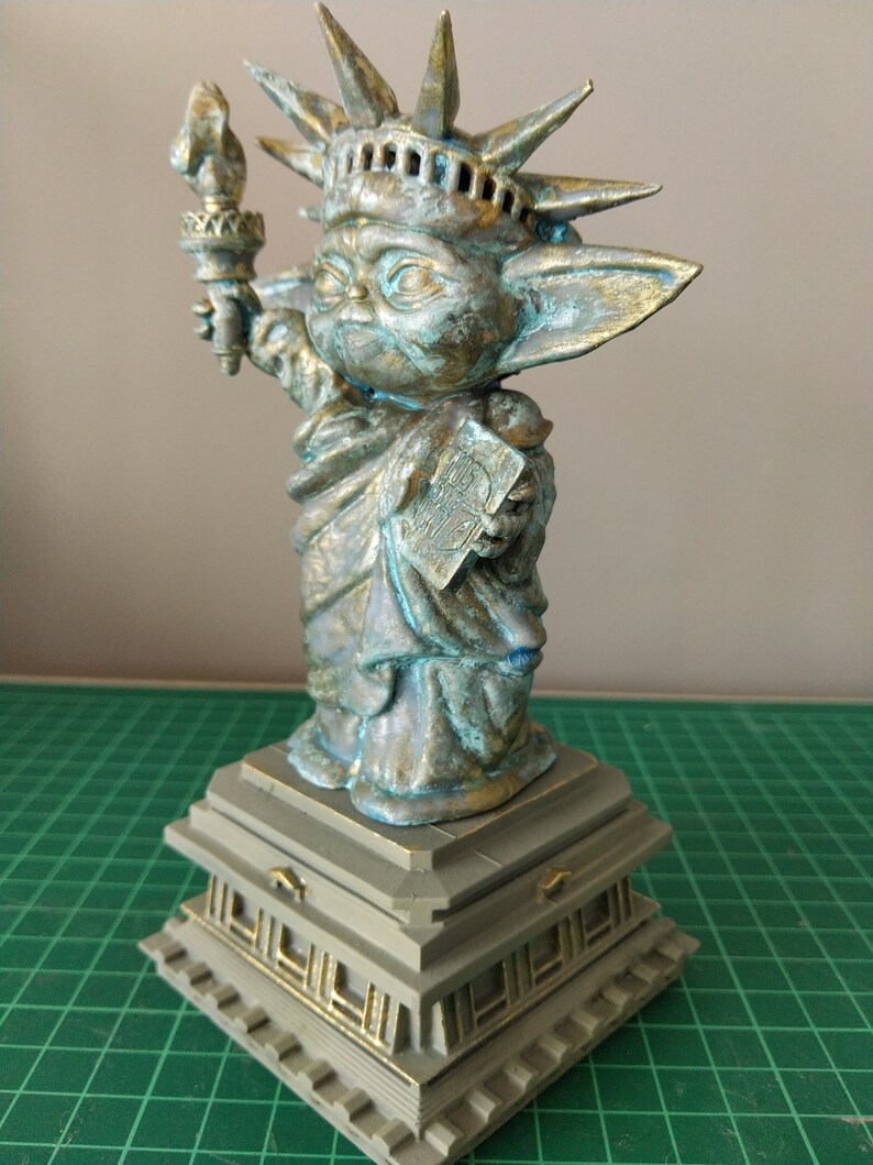 May include: A green and gold statue of the Statue of Liberty, but with the head and body of a Baby Yoda character. The statue is standing on a square base with decorative details.