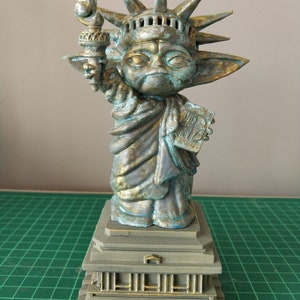 May include: A green and gold statue of the Statue of Liberty, but with the head and ears of a Yoda character. The statue is holding a torch in its right hand and a book in its left hand. The statue is standing on a base with a gold trim.