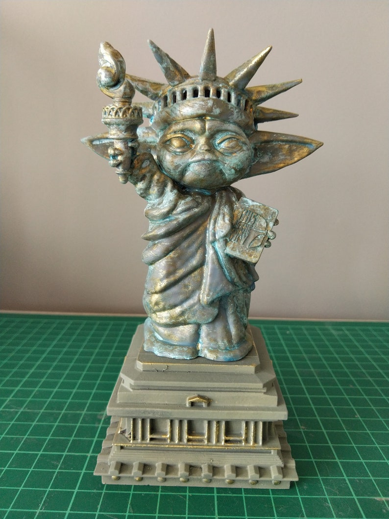 May include: A green and gold statue of the Statue of Liberty, but with the head and ears of a Yoda character. The statue is holding a torch in its right hand and a book in its left hand. The statue is standing on a base with a gold trim.