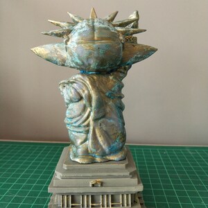 May include: A small, green and gold statue of the Statue of Liberty, with a crown of spikes and a torch in its right hand. The statue is standing on a gray base with a series of steps.
