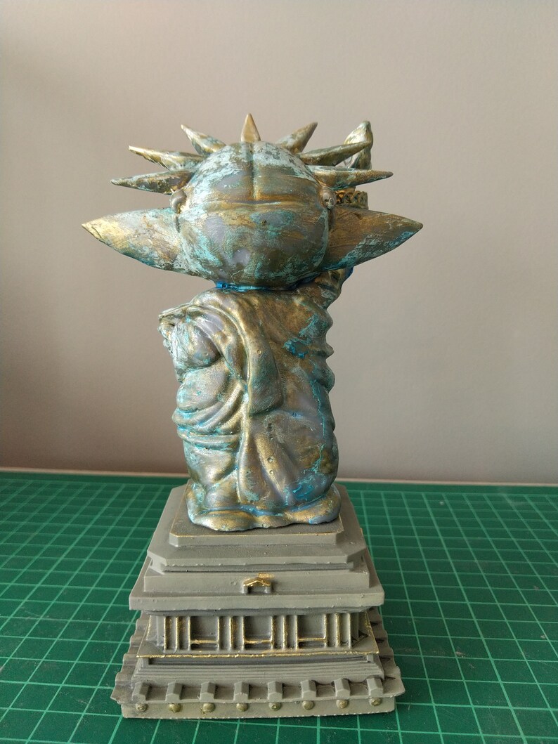 May include: A small, green and gold statue of the Statue of Liberty, with a crown of spikes and a torch in its right hand. The statue is standing on a gray base with a series of steps.