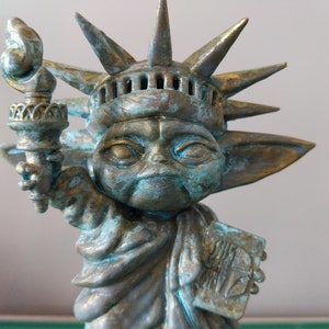 May include: A green and gold statue of a fictional character holding a torch, styled as the Statue of Liberty. The statue has a crown with spikes and a face with large eyes. The statue is holding a torch in its right hand and a book in its left hand. The book has the text "This is the Way" on it.