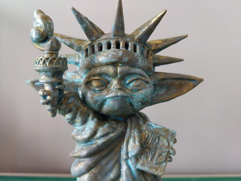 May include: A green and gold statue of a fictional character holding a torch, styled as the Statue of Liberty. The statue has a crown with spikes and a face with large eyes. The statue is holding a torch in its right hand and a book in its left hand. The book has the text "This is the Way" on it.