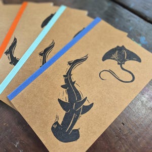 May include: Kraft paper notebooks featuring black sea creature designs. The notebooks have coloured spines in orange, light blue, and blue. Illustrations include a hammerhead shark and a stingray.