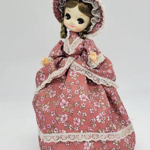 May include: A vintage doll wearing a floral print dress and matching bonnet. The dress is a dusty rose color with a white floral pattern and white lace trim. The doll has a painted face with large eyes and a sweet expression.