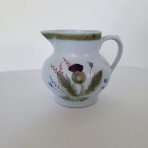 May include: A light blue ceramic pitcher with a handle and spout. The pitcher is decorated with a floral design featuring purple, pink, and blue flowers, and brown leaves. The rim has a brown band.